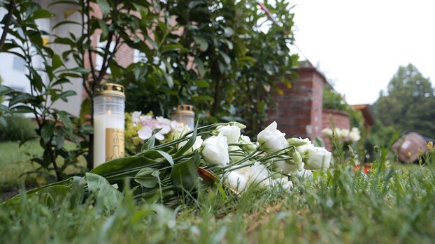 Blumen und Kerzen liegen und stehen im Gras vor einem Backsteingeb&auml;ude. Es ist die Jugendeinrichtung, in der mehrere Menschen am Montag, 29. Juni 2026, durch Sch&uuml;sse gestorben sind.