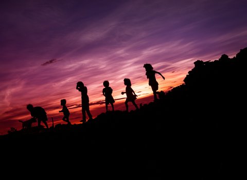 Umrisse von einer Gruppe von Kindern, die vor einem lila-roten Himmel laufen oder spielen und von denen nur die Umrisse zu sehen sind. 