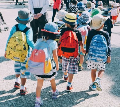 Kindergruppe auf dem Zebrastreifen von hinten fotografiert. Kinder mit bunten Rucks&auml;cken und Sonnenh&uuml;ten &uuml;berqueren die Stra&szlig;e in Begleitung von Erwachsenen. Die Rucks&auml;cke sind in verschiedenen Farben wie Gelb, Rosa, Orange und Blau gestaltet. Sonniger Tag, Alltagsszene im Stra&szlig;enverkehr.