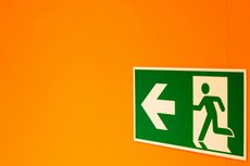 Ein grünes Notausgang-Schild mit Pfeil nach links vor einem orange-farbenen Hintergrund.