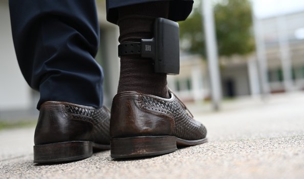 Nahaufnahme von F&uuml;&szlig;en mit elektronischer Fu&szlig;fessel. Zu sehen sind die Unterschenkel einer Person in dunkler Hose und braunen Lederschuhen mit geflochtenem Muster. Am rechten Kn&ouml;chel ist eine schwarze elektronische Fu&szlig;fessel befestigt. Die Person steht auf einer hellen Betonoberfl&auml;che, im unscharfen Hintergrund sind Geb&auml;ude und Gr&uuml;nfl&auml;chen zu erkennen.