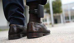Nahaufnahme von Füßen mit elektronischer Fußfessel. Zu sehen sind die Unterschenkel einer Person in dunkler Hose und braunen Lederschuhen mit geflochtenem Muster. Am rechten Knöchel ist eine schwarze elektronische Fußfessel befestigt. Die Person steht auf einer hellen Betonoberfläche, im unscharfen Hintergrund sind Gebäude und Grünflächen zu erkennen.