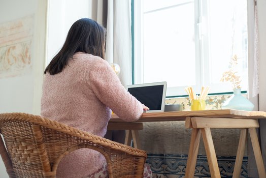 Eine Person mit langen Haaren sitzt an einem Schreibtisch an ihrem Laptop. Der Schreibtisch steht so, dass die Person auch gleichzeitig nach drau&szlig;en sehen kann. 