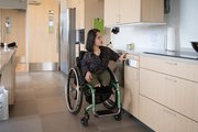 Frau im Rollstuhl in einer hellen, barrierefreien K&uuml;che; sie sitzt am K&uuml;chentresen und greift nach einer Schublade.
