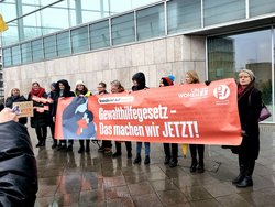 Gruppe von Menschen steht bei einer Kundgebung im Freien und hält ein großes Banner mit der Aufschrift „Gewalthilfegesetz – Das machen wir JETZT!“. Kleinere Schilder mit Aussagen zu Schutz vor Gewalt und Femiziden sind ebenfalls sichtbar. Das Gebäude im Hintergrund hat eine moderne Glasfassade; die Personen sind wetterfest gekleidett.